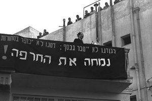 A black and white photo shows Menachem Begin standing on a blacony with a banner in Hebrew hanging on the side.