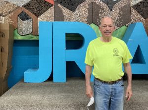 A white man in jeans and a neon yellow short stands in front of large block letters that read "JRA".