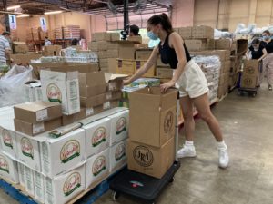 A white woman in a tank top, shorts and mask loaded dry foods into boxes stacked on a cart.