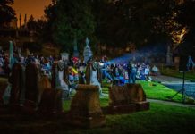 A projector illuminates gravestonesa nd a gaggle of guests watching a movie on the cemetery grass.