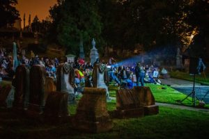 A projector illuminates gravestonesa nd a gaggle of guests watching a movie on the cemetery grass.