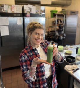 Rachel Klen is a white woman with blonde hair wearing a flannel. She is displaying a botton of green sauce and has her tattooed arms exposed.