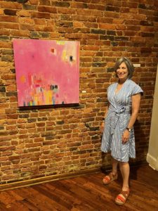 A white woman stands next to a painting hung on an exposed brick wall.