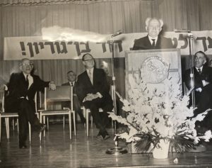 A black and white photo shows David Ben-Gurion speaking behind a podium on a stage iwht several other men in suits sitting on the stage.