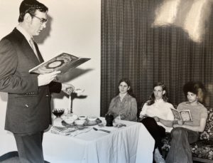 A black and white photo shows a rabbi leading a seder in front of three young women sitting at a table.