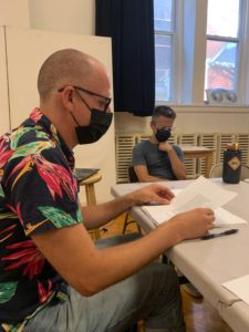 A bald white man in a color, short=sleeved button up shirt is wearing a mask and sitting at a table reading from a script.