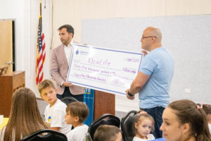 Brian Gralnick, a white man with short hair, hands a giant check to Andre Krug, a white, bald man wearing a blue t-shirt.