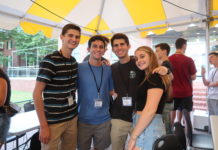 A group of students in shorts and t-shirts stand under a tent with their arms wrapped around each other.
