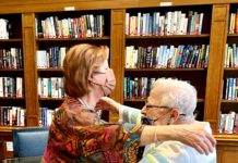 Two women embrace in a hug. They are standing in front of a book shelf.