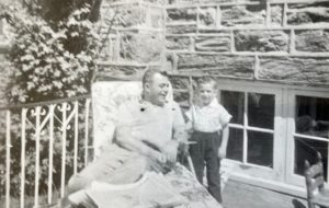 In a black and white photo, Drew Trachtenberg stands next to his father, who is lounging in a chair outside.