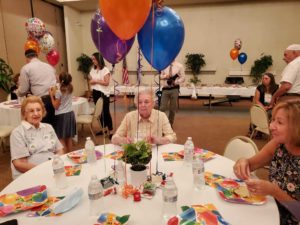 Survivors gather around a circular dining table decked with balloons and festive plates and decorations.
