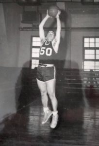 Myron Rosenbaum is wearing a basketball uniform and is jumping in the air, hands outstretched over his head.