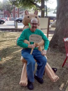 Myron Rosenbaum is sitting on a large inflatable animal and holding a sign that reads, "Need a hug? Take a seat!"