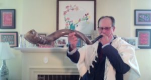 Phill Goldberg is a white man in a suit and tallit; he is holding up a long shofar and blowing into it.