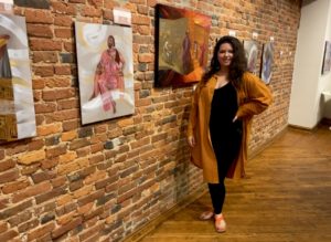 Sasha Zeiger is a white woman with long, curly hair wearing a long, orange cardigan standing in front of an exposed brick wall, which displays her paintings.