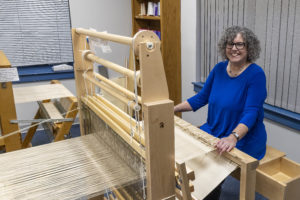 Ilene Cetlin Lipow is a white woman with curly salt-and-pepper hair wearing a blue dress and sitting behind a large wooden loom.