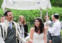 Walking down the aisle at an outdoor venue, Andrew Davies, wearing a suit and tallit, holdsthe hand of Molly Wernick, wearing a white dress