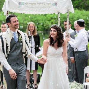 Walking down the aisle at an outdoor venue, Andrew Davies, wearing a suit and tallit, holdsthe hand of Molly Wernick, wearing a white dress