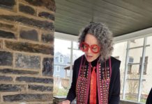 Louie Asher, a white woman with neatly parted grey hair, is wearing bright red classes and tie and is reading from the megillah at a table on a porch.