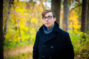 Ari Sussman is a white man wearing a winter coat and scarf and thick-rimmed glasses. His brown hair is swooped to the side.