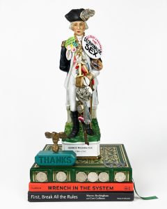 A statue of George Washington is adorned with several pins and buttons draping across his torso. He is perched on several old books.