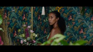 A black woman stands in front of a colorful wall and amid a room filled with flowers.