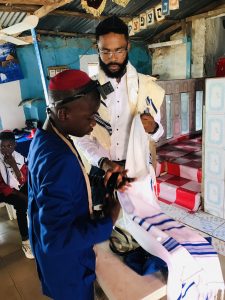 Moshe Hezekiah is a Black man with a short beard and glasses wearing a tallit and helping a young Nigerian Jewish boy wrap tefillin.