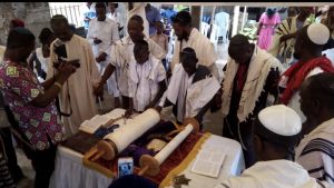 A group of Nigerian Jews wearing tallitot huddle around the davening stand with a Torah rested on it.