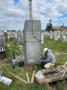 A crane lifts a large gravestone while a man kneels and guides it back onto the ground.