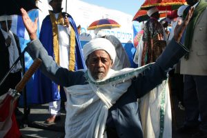 An Ethiopian man in a white hat and robes kneels with his arms up and palms open.