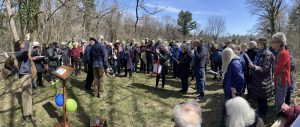 Congregants stand in a large field surrounded by trees.