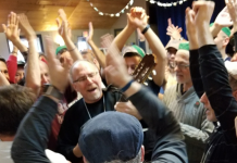 Rabbi Shawn Zevit is a white man with grey hair and goatee strumming a guitar and singing, surrounded by man dancing.