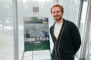 Zeke Winitsky is a white man with a red beard and red curly hair. He is wearing a cardigan and standing next to his movie poster.