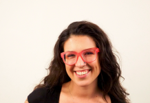 Emma Chasen has long, dark hair and a sleeve of tattoos. She is wearing bright red glasses and smiling at the camera.