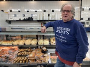 Russ Cowan is a white man with glasses wearing a "Radin's" sweatshirt, learning on a glass deli case willing with cookies and cakes.