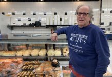 Russ Cowan is a white man with glasses wearing a "Radin's" sweatshirt, learning on a glass deli case willing with cookies and cakes.