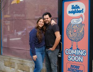 Mike and Alexandra Cohen stand with their arms wrapped around each other outside of their store with a "Coming soon" sign with a bagel on it.