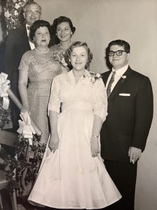 Sybil Klein, wearing a white dress, stands next to Robert Klein, wearing a suit and white pocket square, at the bottom of a set of stairs, with family posing behind them.