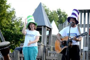 Jenny and David Heitler-Klevans are performing outside on stage, both wearing tall, Cat in the Hat-like hats.