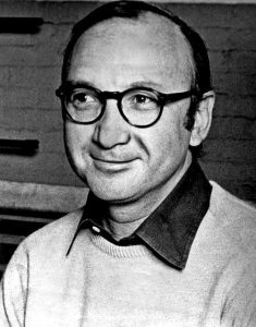 Neil Simon is a white man with thick-rimmed glasses wearing a sweater and collared shirt, smiling and looking to the left.