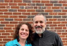 Jenny and David Heitler-Klevans are standing outside in front of a brick wall with their arms wrapped around each other.