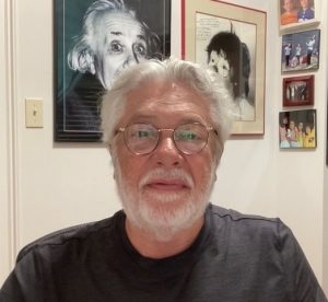 George Trajtenberg, with white hair and beard, glasses and wearing a black shirt, is sitting in front of some framed pcitures.