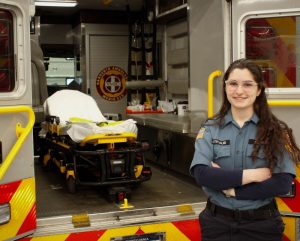 Devora Weintraub is a young white woman wearing a blue EMT uniform standing in front of an open ambulance.