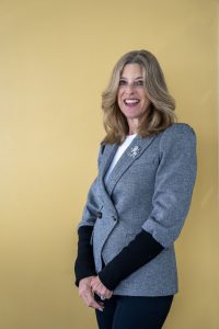 Gail Norry is a white woman with long, wavy blonde hair wearing a grey suit and standing smiling on front of a yellow background.