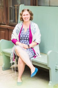 Elyssa Cherney is a white woman with shoulder-length blonde hair wearing a patterned dress and tallit and sitting on a green bench with her legs crossed.