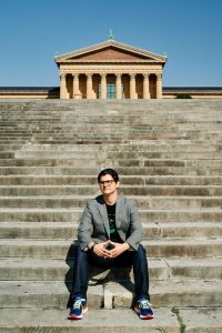 Paul Farber is wearing a suit jacket, jeans and sneakers and is sitting on the PMA steps, the museum looming in the background.