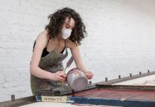 Gavi Weitzman is wearing an apron and mask and pouring paint onto a screen print.