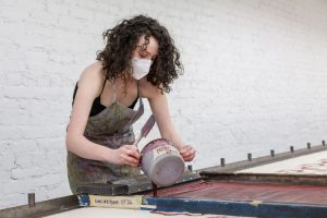 Gavi Weitzman is wearing an apron and mask and pouring paint onto a screen print.