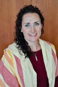 Kami Knapp Schechter is a white woman with long, curly brown hair and blue eyes draped in a pink, orange and white tallit gadol.