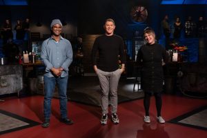 Three chefs stand side-by-side in a dimly-lit studio kitchen.